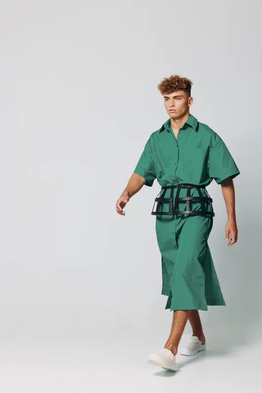 Young man walking in green oversized shirt and matching wide pants with white sneakers on plain background. Fashion model with curly hair showcasing modern casual outfit in studio.