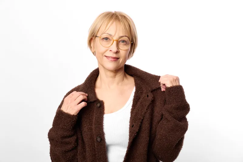 Woman elderly glasses portrait coat fashion casual smiling, studio closeup of mature female adjusting brown jacket, relaxed confident mood on clean white background.