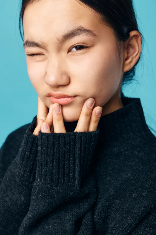 Young woman with dark hair wearing black sweater winks and touches her chin in closeup portrait on blue background. Facial expression, casual and thoughtful mood with natural light.