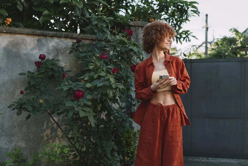 Cheerful woman outdoors, wearing rust outfit, holding smartphone, relaxed pose, sunlit garden with roses, natural colors, soft breeze, casual lifestyle moment, vivid light, everyday scenery