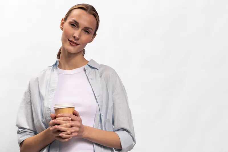 Young woman in casual clothes holds coffee cup, smiles gently, standing against white background. Concept for relaxation, morning routine, and lifestyle.