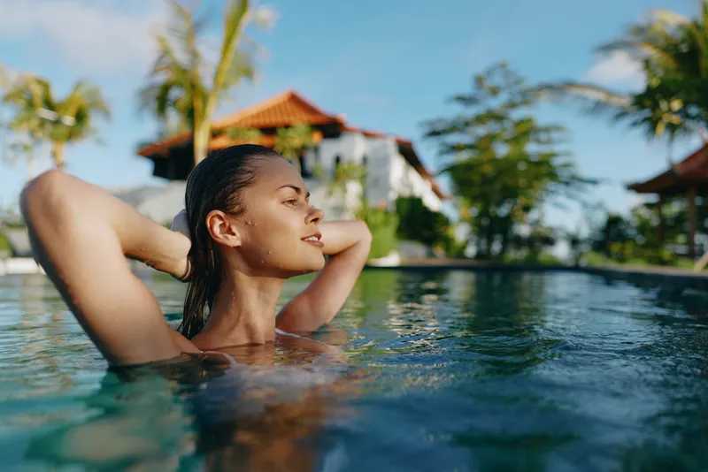 Relaxed woman in a tropical pool enjoys a calm moment, water ripples around her, warm sun, serene mood, vacation vibe, outdoor retreat with palm trees and blue sky.