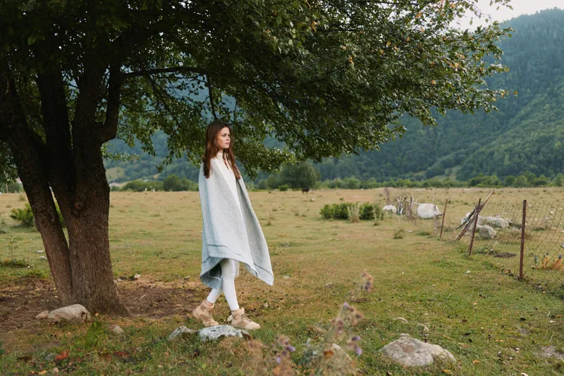 Woman in a light shawl stands near a large tree in a tranquil countryside, presenting a calm lifestyle moment, connection with nature, and a relaxed casual pose