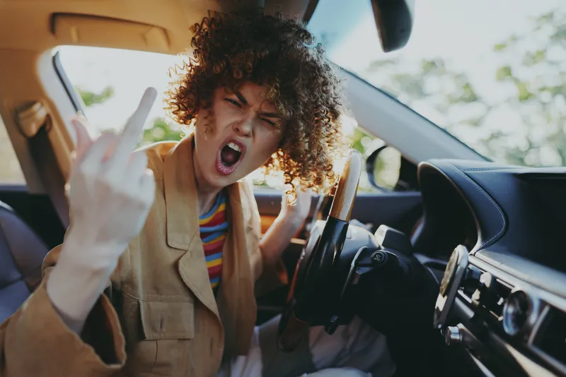 A woman with curly hair in a tan jacket sits inside a car, flashing a bold gesture and a strong attitude that signals rebellious lifestyle. Retro film color mood enhances the edgy, cinematic vibe.