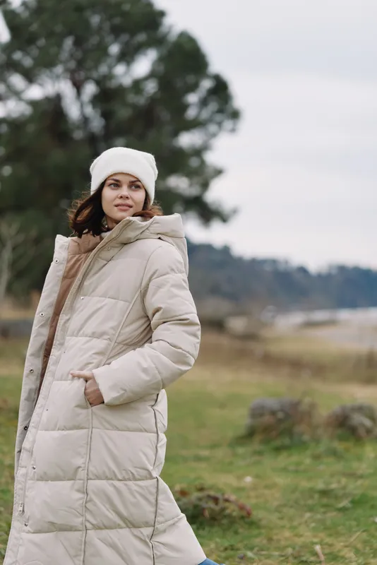 Woman in a thick winter coat and beanie stands in an open field with trees, calm winter scenery, fashion and outdoor comfort for cold season portraits
