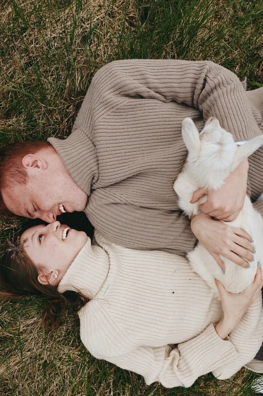 A couple lounges on a grassy field, embracing a white goat kid in a warm, wholesome lifestyle moment, expressing affection and calm energy in an outdoor setting, with gentle laughter, casual