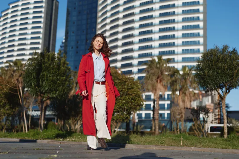 Young woman walking outdoors in a red coat and white pants with modern buildings and trees in the background on a sunny day. Urban lifestyle and fashion concept.