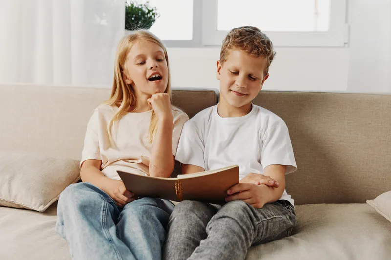 Two children sitting on a beige sofa reading a book together, enjoying a quiet moment in a bright living room. The girl is singing while the boy follows the story attentively.