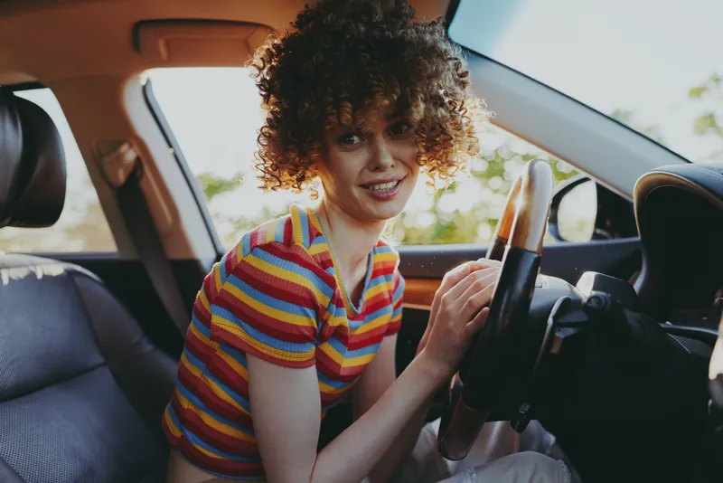 Smiling woman with curly hair wearing a striped shirt sits behind the wheel of a car, creating a casual lifestyle moment with warm retro tones and natural daylight. Relaxed mood.