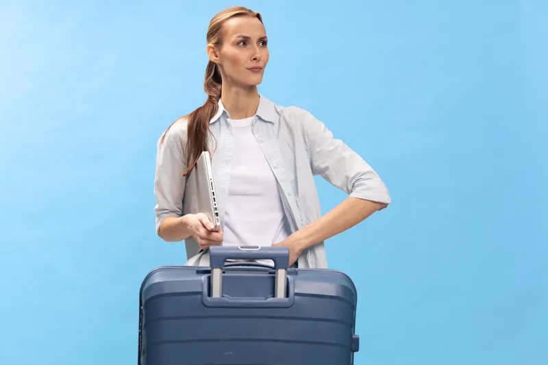 Young woman with suitcase and laptop stands on blue background, looking confident and prepared. Concept for travel, business trip, and modern lifestyle with technology.
