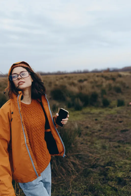 Woman outdoors lifestyle in countryside, wearing jacket and glasses, holding smartphone, relaxed pose, cloudy daylight, looking ahead, soft wind, natural colors, grassy field today