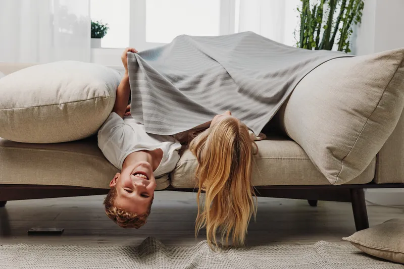 Happy young couple lying upside down on sofa under blanket in cozy bright living room. Playful mood, casual home leisure, and intimate relationship moment with natural light and soft decor.