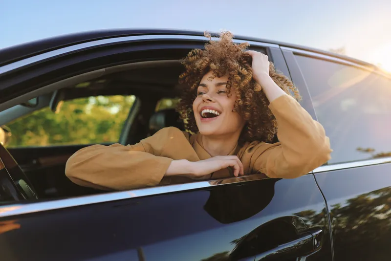 Joyful woman leans out of a car window, smiling at the sunny day, capturing a candid travel moment with retro film color and warm hues, conveying carefree lifestyle and outdoor freedom