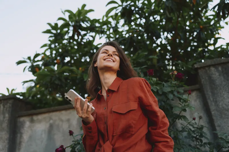 Cheerful woman outdoor, smiling, holding a phone, casual red shirt, sunlit moment, relaxed posture, natural colors, soft breeze, lush greenery background in a bright setting
