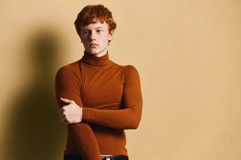 Young man with red hair wearing orange turtleneck sweater standing against beige background with serious expression and folded arms for portrait and fashion style.