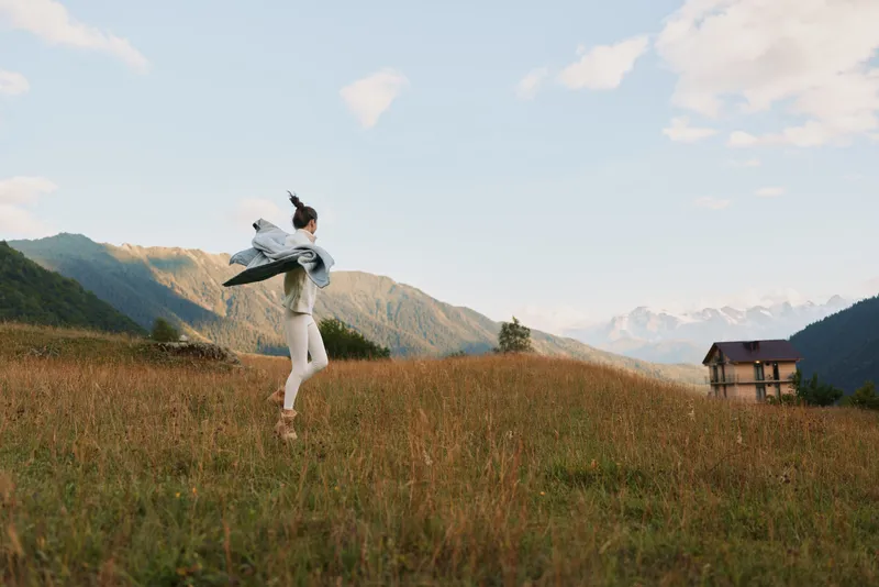 Woman in white dress dances in a sunlit meadow with mountains in the background, showcasing carefree lifestyle and connection with nature, isolated on expansive field.