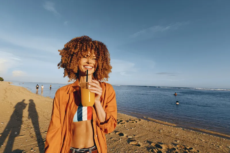 Beach woman curly hair smile ocean shoreline sunlight summer outdoors, cheerful tourist standing on sandy coast, smiling into camera, warm breeze moving hair, swimmers under blue sky