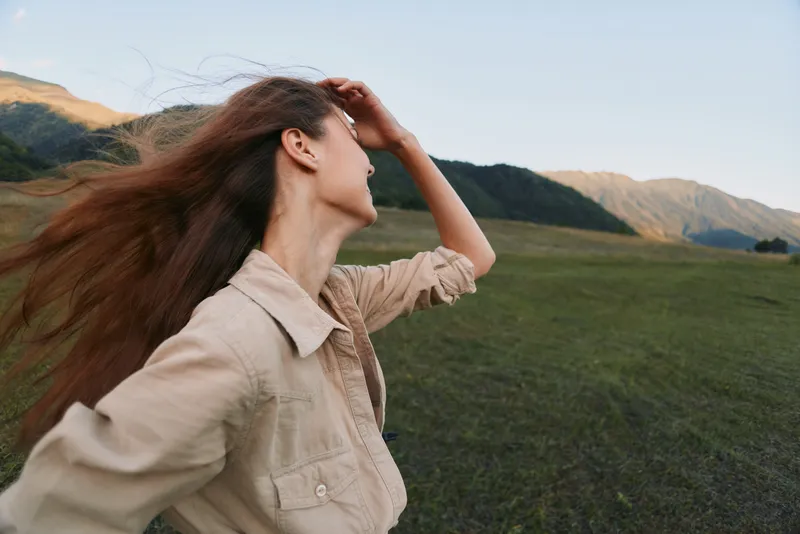 Woman outdoors in a wind swept field, mountains in the background, relaxed lifestyle portrait, sunlit natural colors, breezy moment in open nature