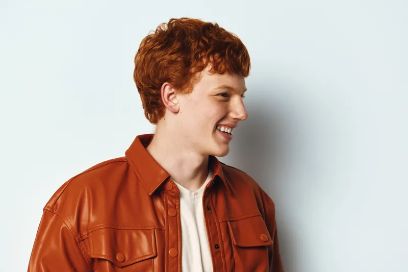 Young man with red hair wearing orange leather jacket and white shirt smiling looking to side on light background in casual style portrait photography.