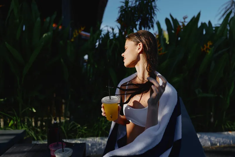Casual lifestyle scene of a woman by a poolside, sipping juice through a straw, wrapped in a light shawl, gazing to the side as tropical plants frame a sunny moment.