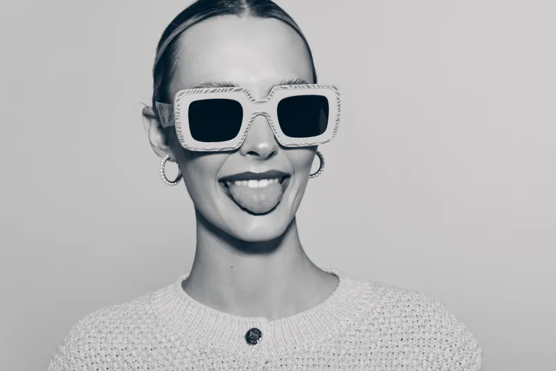 Young white woman with fair skin in oversized sunglasses and white sweater shows playful expression sticking out tongue in studio. Concept of fun and fashion.
