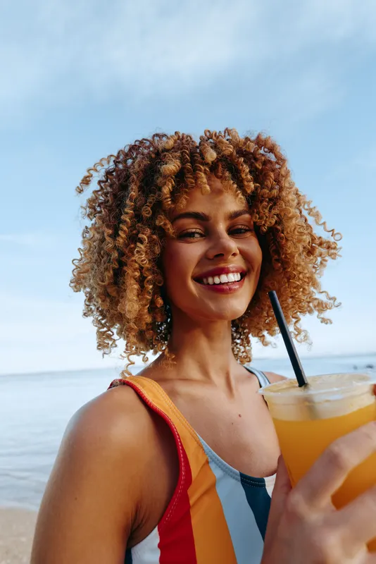 Young woman with curly hair smiles happily while holding a cold drink at the beach on a sunny day, enjoying a refreshing moment by the sea.