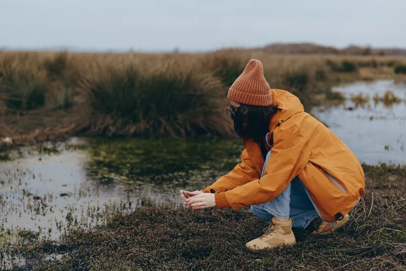 Woman lifestyle outdoors by river and pond during autumn winter, wearing orange coat and knitted hat, crouching on muddy shore with boots, hands reaching forward, calm nature landscape