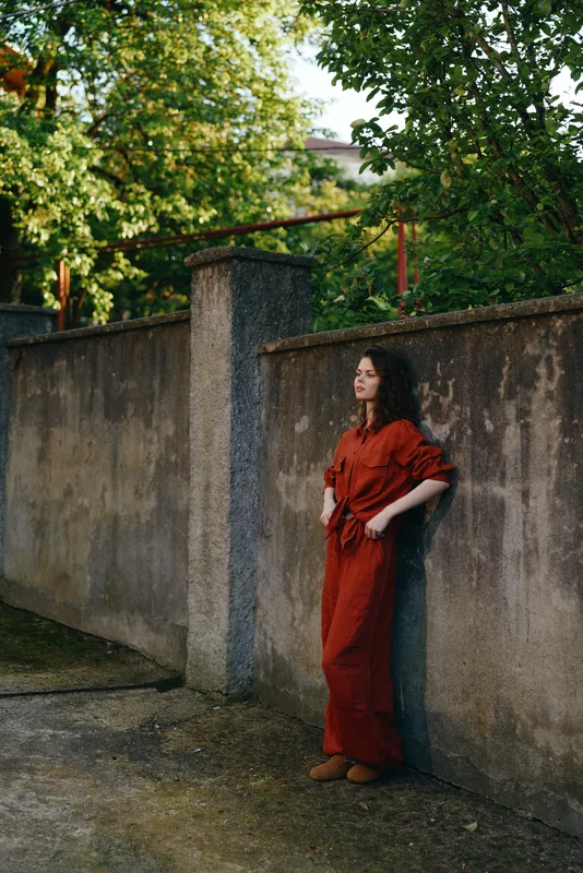 Woman in a red jumpsuit leaning against a concrete wall in a sunny outdoor setting, showcasing casual chic style, relaxed pose, and urban nature harmony