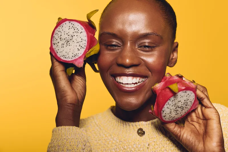 Smiling black African woman with dark skin holds two halves of dragon fruit near face on yellow background. Concept of healthy lifestyle, tropical fruit, natural beauty, and positive mood.