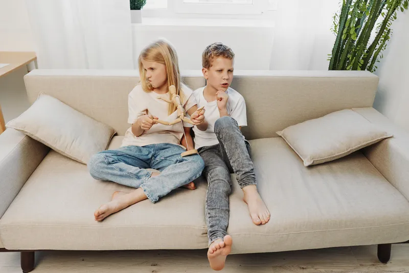 Two children with serious expressions sit apart on a beige sofa in a bright living room, showing tension and conflict between siblings indoors.