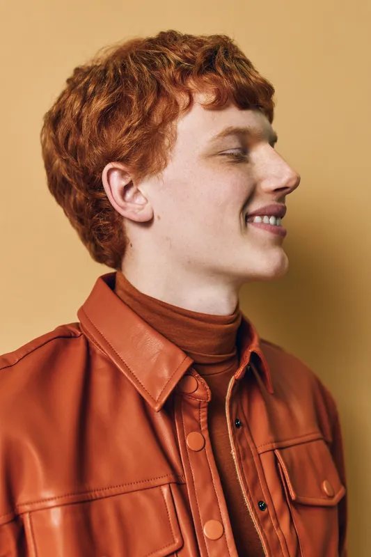 Young man with red hair wearing a brown leather jacket and turtleneck smiling and looking sideways on beige background, casual style and profile portrait for fashion concept.