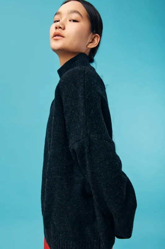 Portrait of young woman with dark hair wearing black sweater posing against blue background. Studio shot with fashion style and confident attitude for casual clothing and beauty concept.