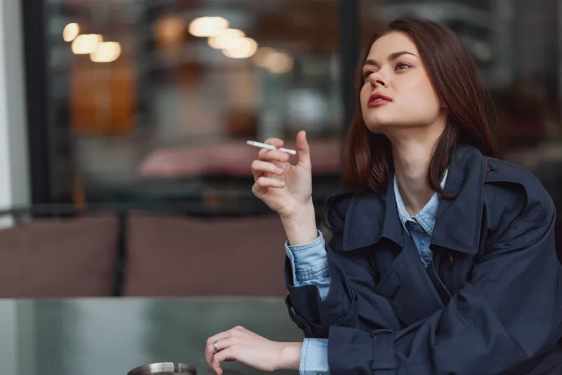 Young woman in dark coat smokes cigarette with thoughtful expression sitting outdoors in urban cafe, blurred background with warm lights, casual lifestyle concept.