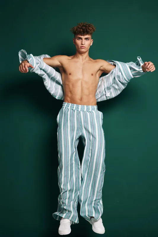 Shirtless young man with curly hair wearing striped pants and holding open a matching shirt posing confidently in front of dark green background in studio portrait.