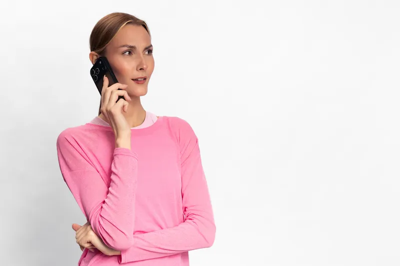 Young woman in pink sweater talking on smartphone with thoughtful expression on white background. Concept of communication, modern lifestyle, and casual technology use.
