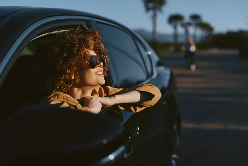 Woman with curly hair leans from a car window, sunglasses on, enjoying sunlit street in retro film color aesthetic, candid lifestyle moment, joyful and relaxed.