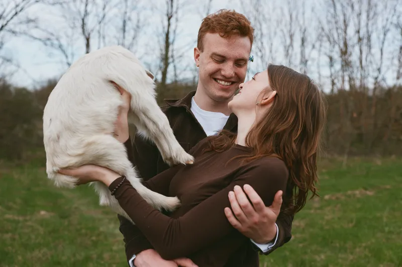 A couple outdoors, lifestyle scene, affection for a goat kid, sharing a warm laugh as they cradle the playful animal, highlighting companionship, happiness, and nature in a candid outdoor moment.