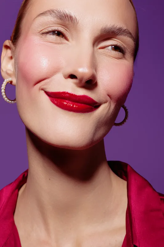 Close-up of smiling woman with red lipstick and hoop earrings on purple background. Concept of beauty, makeup, fashion, and confident female portrait with vibrant colors and studio lighting.