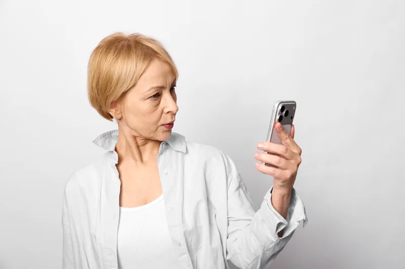 Elderly woman holding a smartphone for selfie on a white background, surprised expression and gaze. Technology, communication, and digital life concept with soft light.