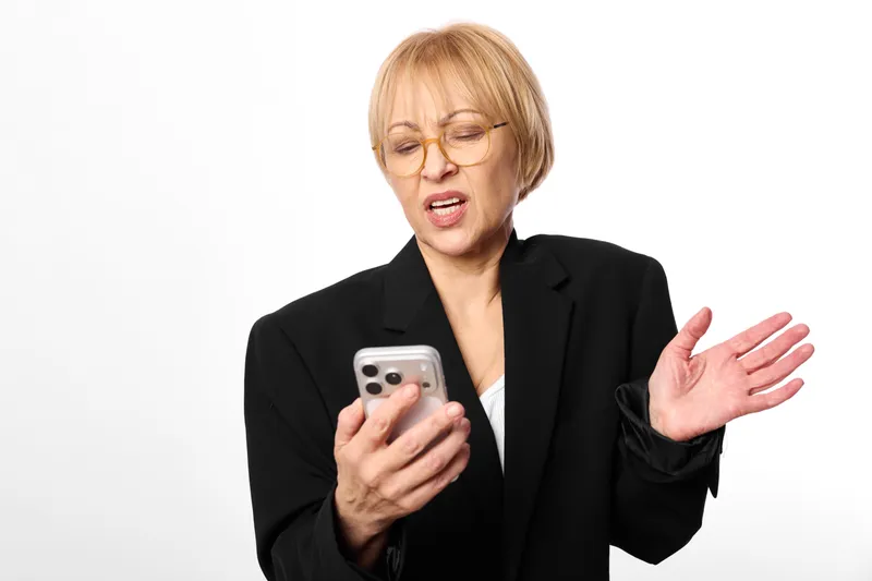 Elderly woman with glasses holding a smartphone, looking surprised. Studio portrait on white background, black blazer, one hand raised. Communication and technology concept.