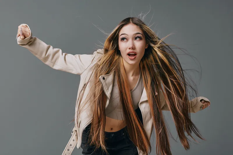Young woman with long hair wearing beige jacket and crop top posing dynamically on gray background for fashion and lifestyle portrait photography with expressive face and movement.