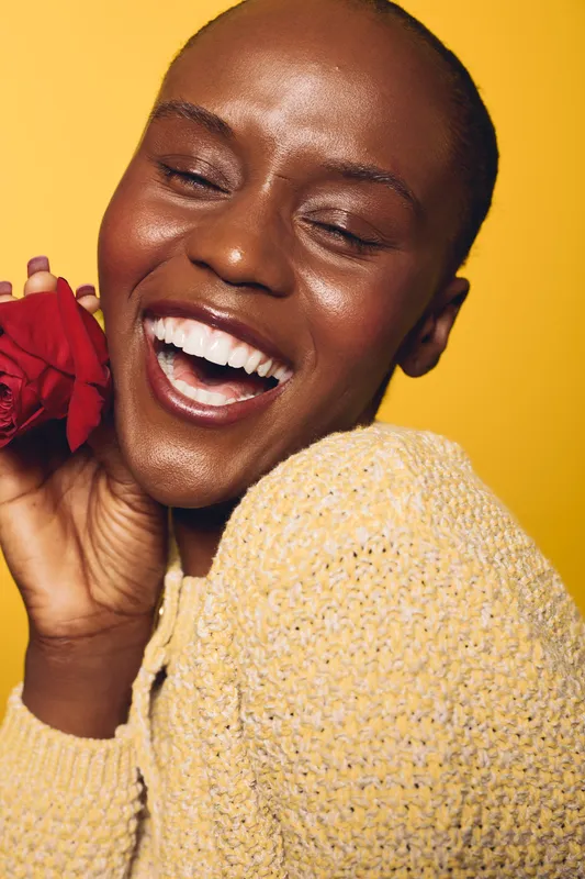 Happy African American woman with dark skin laughs joyfully holding red rose near face. Yellow background and knitted sweater create warm and vibrant studio portrait concept of beauty and happiness.