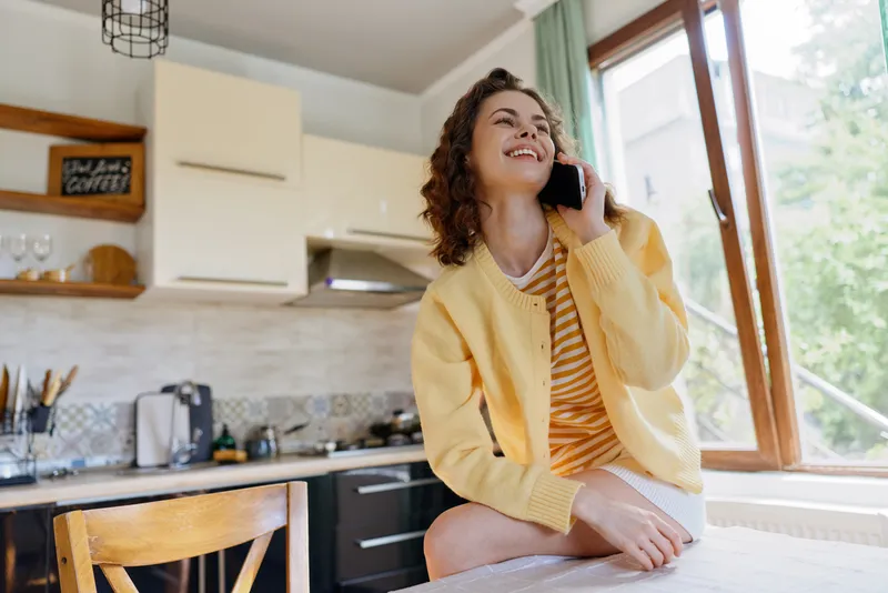 Casual woman at home in a bright kitchen talks on the phone smiling, wearing a yellow cardigan and striped top, creating a warm everyday lifestyle mood in a neat interior