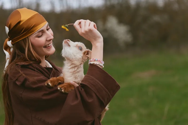 A smiling woman in a brown cloak and orange scarf holds a goat kid close, presenting a yellow leaf. Outdoor lifestyle scene highlighting playful bond and gentle interaction with animals.