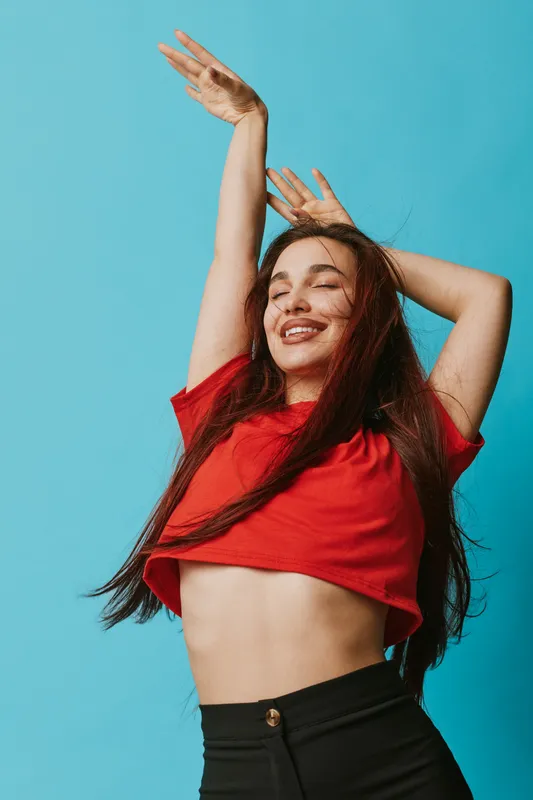 Young woman with long hair and red shirt smiling with eyes closed and hands raised against blue background. Happy girl enjoying freedom and positive energy with casual style.