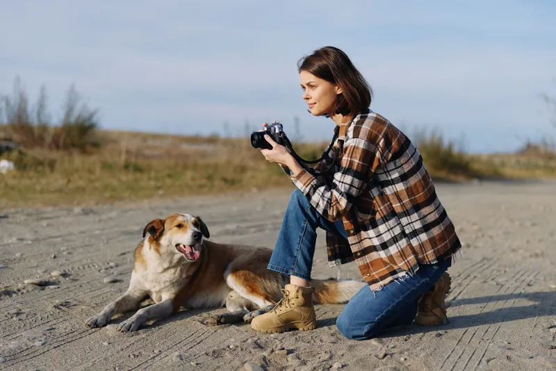 woman photographer kneels on sandy outdoor terrain with a dog beside her holding a camera and focusing on subjects in the open landscape under a clear sky