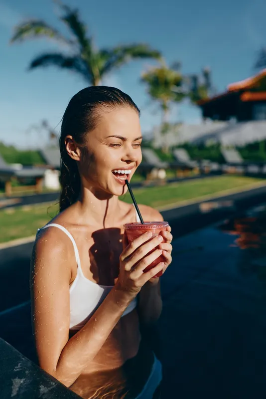 Happy woman in a white swimsuit enjoys a refreshing smoothie by a sunny pool, palm trees in the background, capturing a relaxed tropical summer lifestyle and outdoor leisure.
