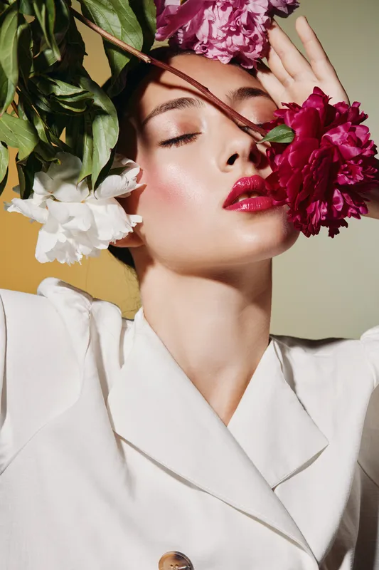 Close-up portrait of a young woman with flawless makeup and glowing skin surrounded by vibrant flowers. Artistic beauty and fashion creativity on a soft pastel background.