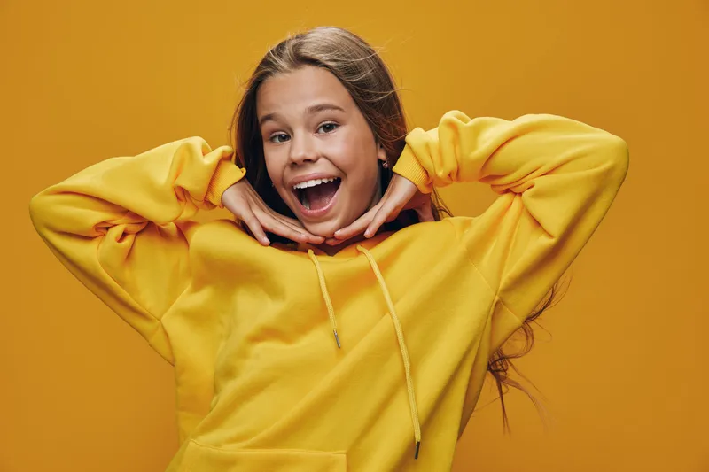 Girl smiling and posing with raised hands near face wearing yellow hoodie against plain background. Young female expressing happiness and energy with casual outfit.