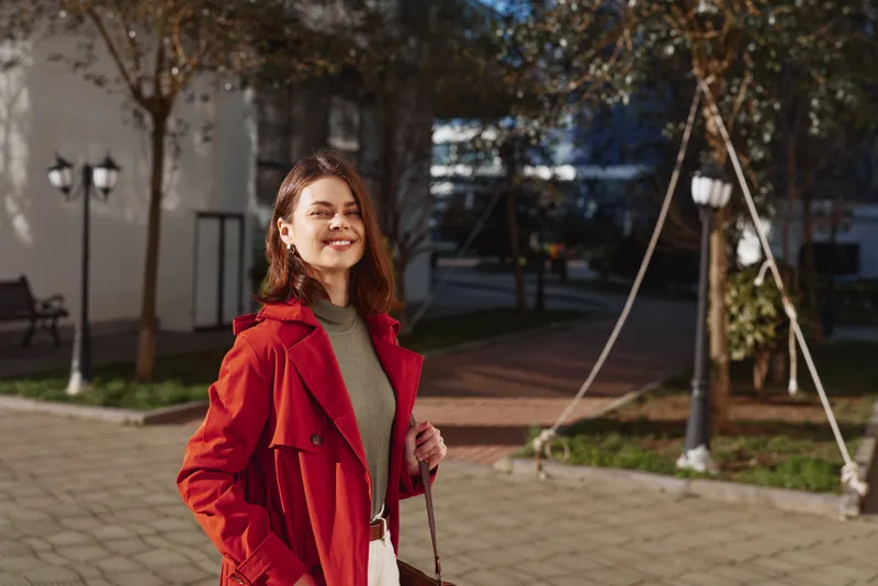 Young woman in a red coat outdoors, smiling under afternoon light, walking in a sunny plaza with trees and modern buildings, a soft breeze rustling her hair for a candid lifestyle moment